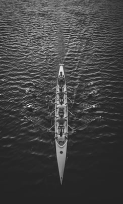 A dramatic bird's eye view of a rowing team riding against the waves of a huge body of water — in black and white. Despite the vastness of the open water, the team pushes forward in unison.