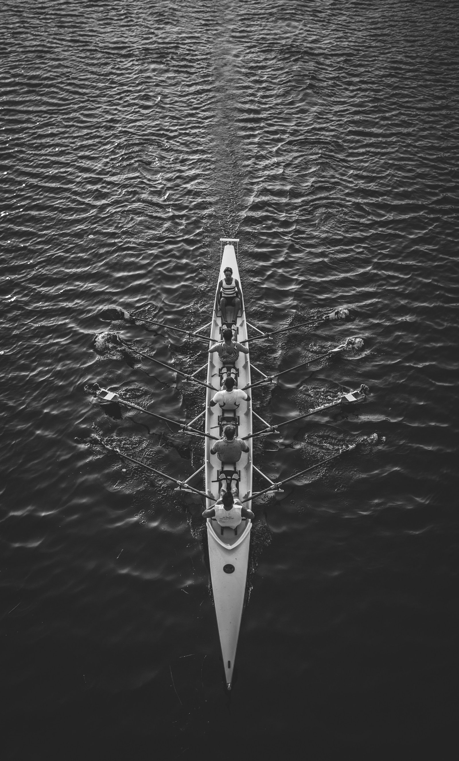 A dramatic bird's eye view of a rowing team riding against the waves of a huge body of water — in black and white. Despite the vastness of the open water, the team pushes forward in unison.
