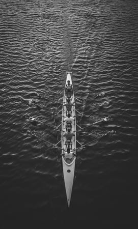 A dramatic bird's eye view of a rowing team riding against the waves of a huge body of water — in black and white. Despite the vastness of the open water, the team pushes forward in unison.